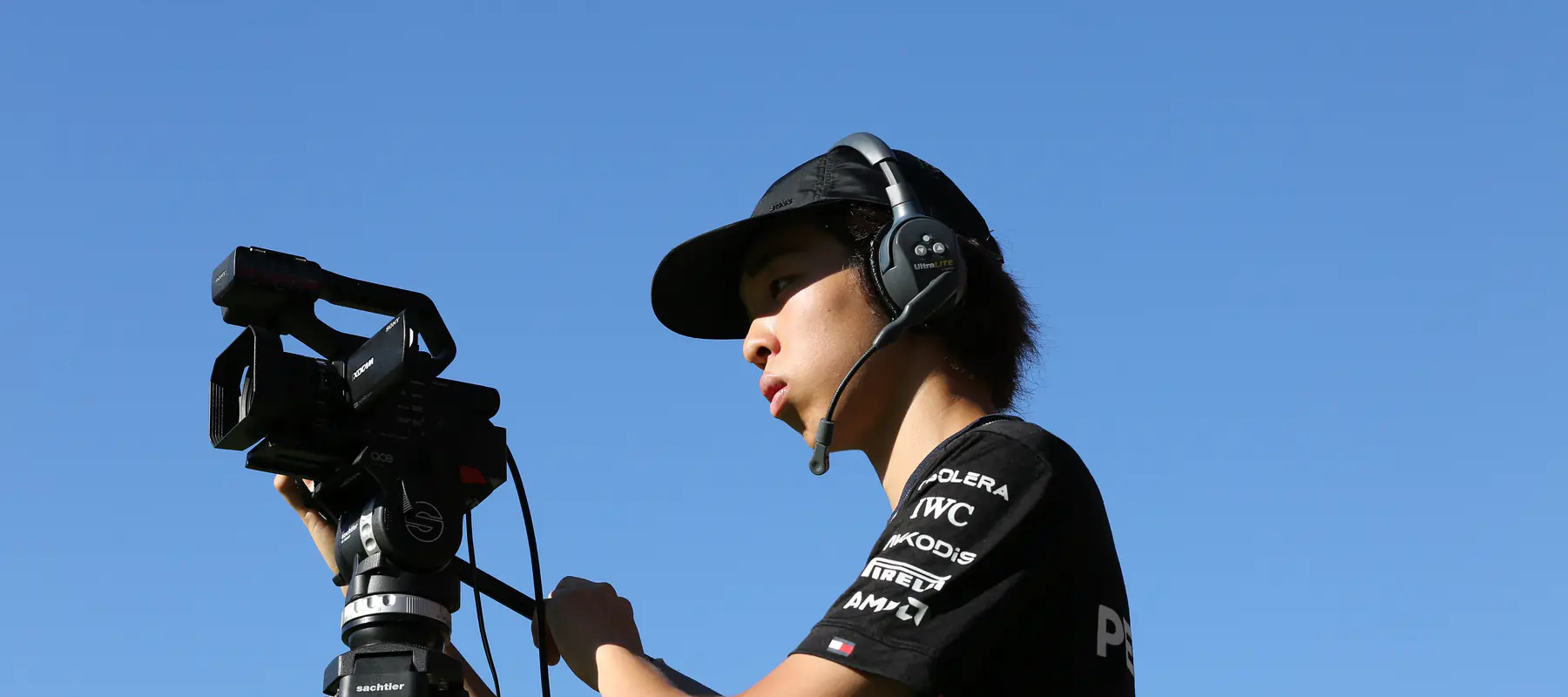 A Media Club student operating a broadcast camera and wearing a production headset outdoors